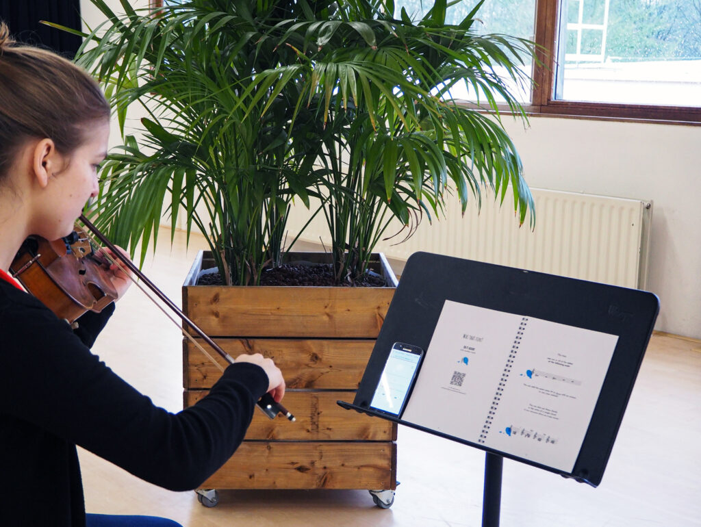 A photo of a woman playing the violin in front of a practice book, with a mobile phone placed next to it.