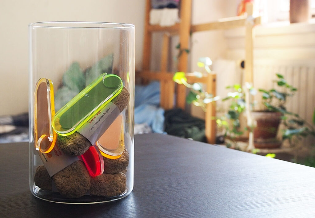 Photo of a glass jar placed on a table in a brightly lit room, containing colourful pill-shaped plastics.