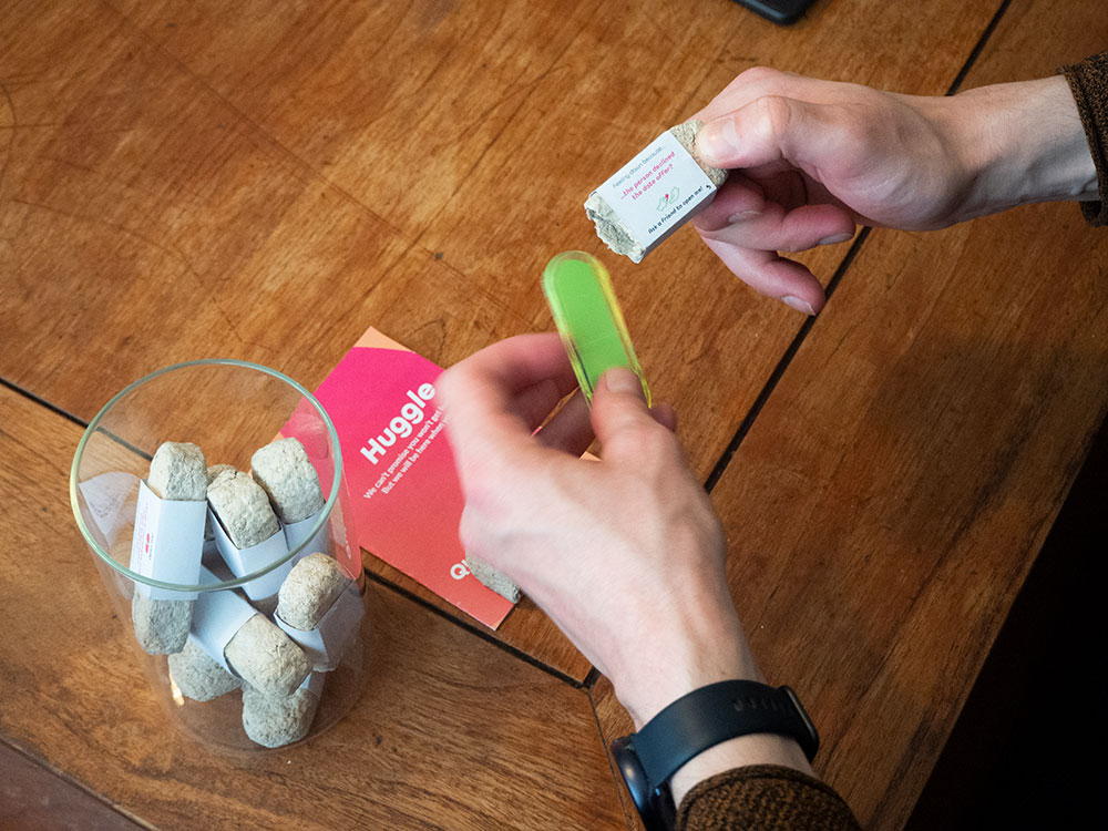A photo of a pair of hands breaking open the mycelium packaging to reveal a colourful, green, pill-shaped plastic object. 